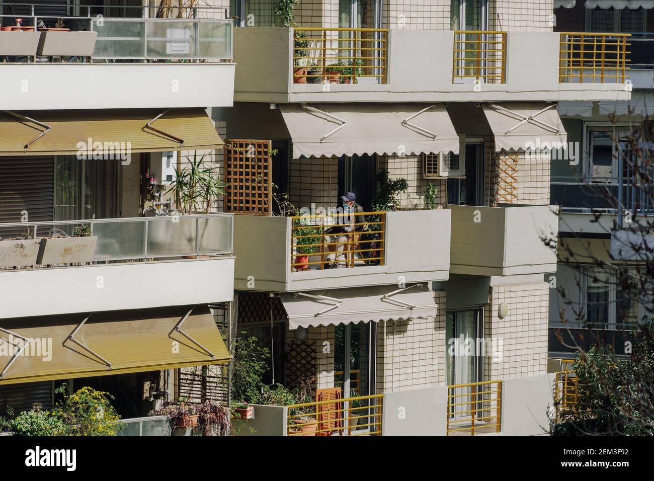 Vecchio uomo che indossa una maschera chirurgica covid-19 al suo balcone. Giorno soleggiato vista di una persona non identificata con coronavirus viso protezione seduta sulla sua veranda in un ambiente urbano città a Salonicco, Grecia. Foto Stock