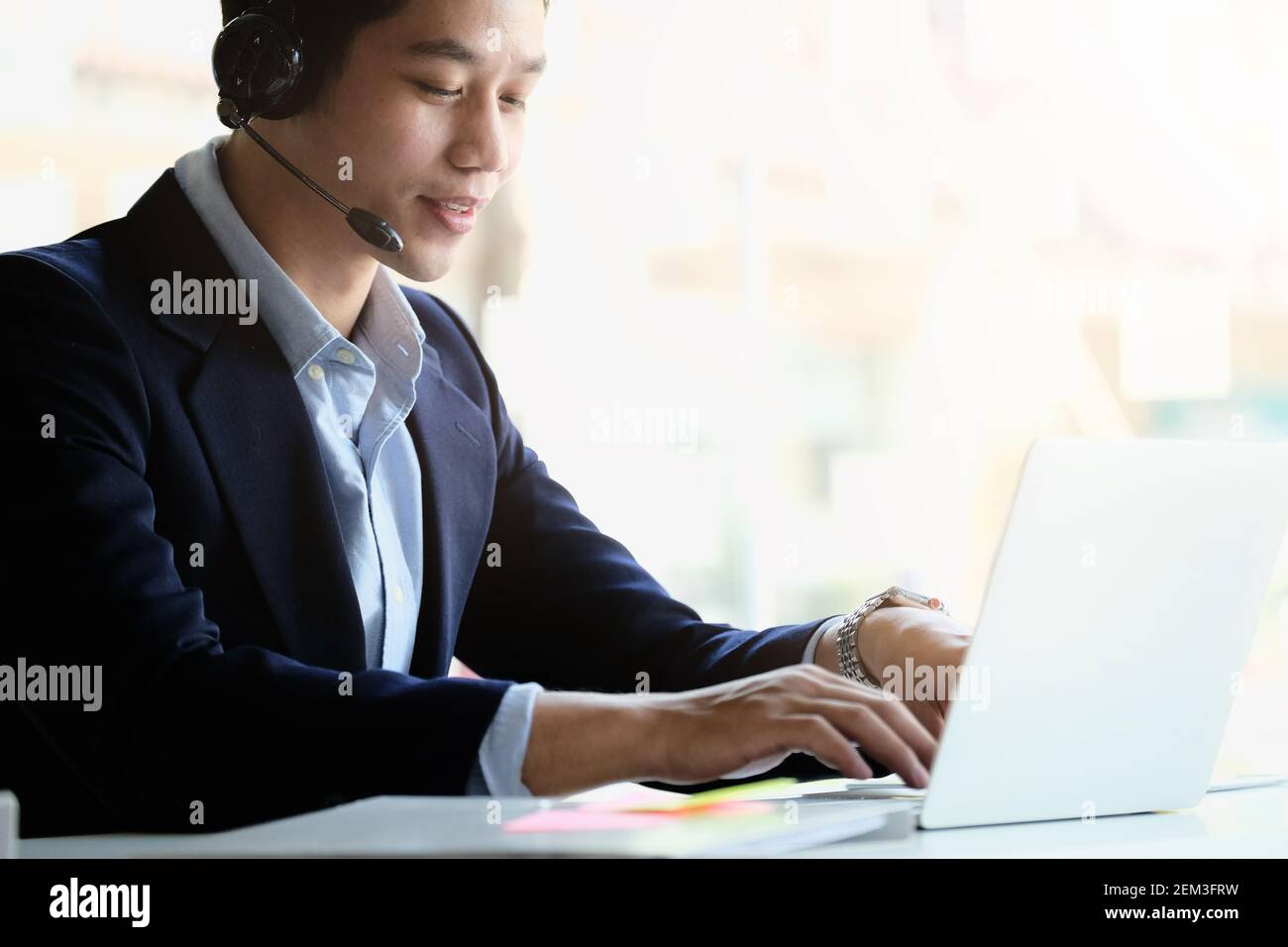 Uomo d'affari asiatico in cuffia che guarda il laptop, guarda un buon webinar, consulente del cliente, imparare a distanza lingue, prendere appunti, uomo felice Foto Stock