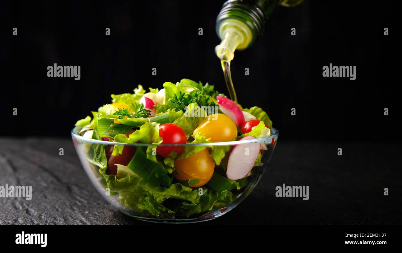 Olio d'oliva versato dalla bottiglia sull'insalata di verdure su nero sfondo Foto Stock