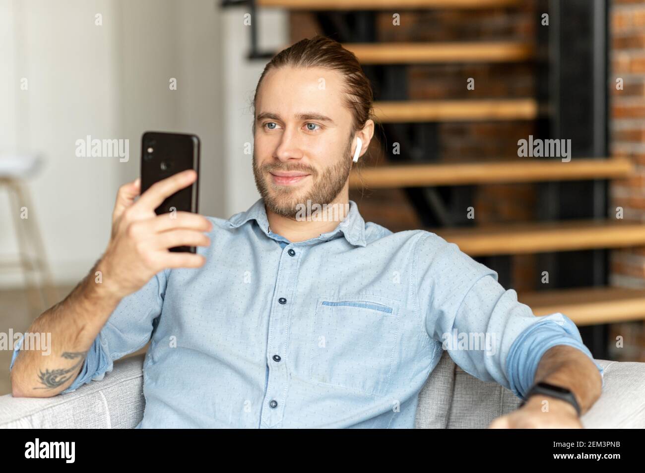 L'uomo europeo con i capelli tirati e la barba sta guardando sul telefono e sorridendo, seduto con gli auricolari orologio fitness nero sul polso, guardando Foto Stock