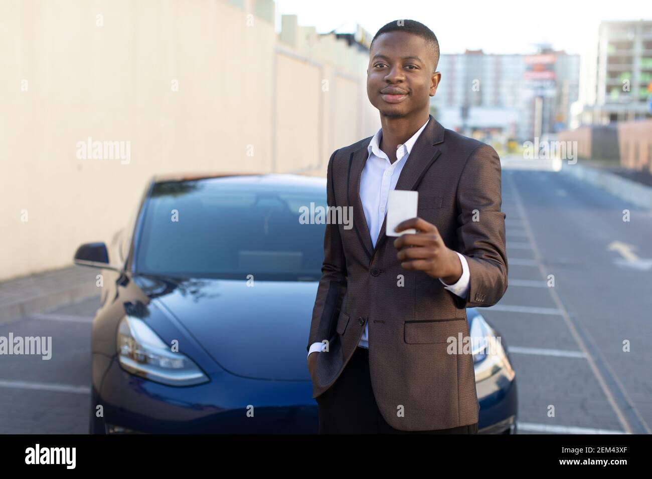 Ritratto di un giovane bell'uomo d'affari afroamericano o manager delle vendite, in piedi con una chiave di carta da una nuova auto elettrica all'aperto presso la concessionaria Foto Stock