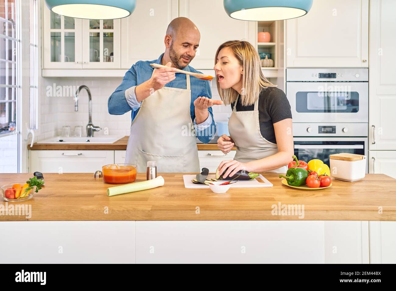 Una giovane coppia felice sta gustando e preparando un pasto sano nella loro cucina. Prepara la salsa di pomodoro e la offre a lei. Concetto di cibo sano Foto Stock