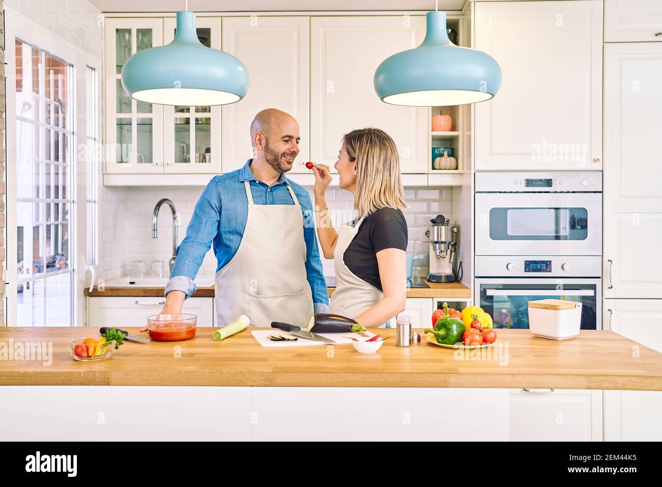 Una giovane coppia felice sta gustando e preparando un pasto sano nella loro cucina. Gli offre un pomodoro. Foto Stock