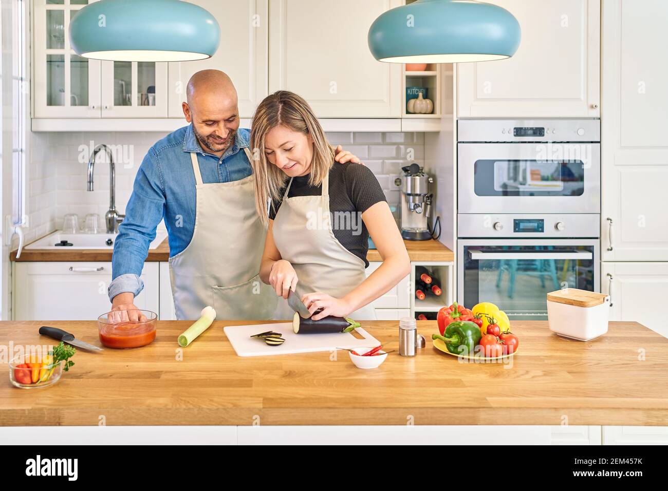 Una giovane coppia felice sta gustando e preparando un pasto sano nella loro cucina. Sta tagliando le verdure mentre la guarda Foto Stock