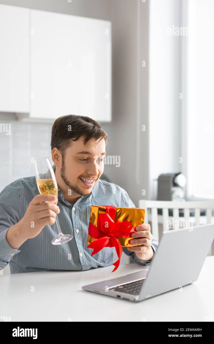 Foto verticale di un uomo con un bicchiere di vino e un giftbox davanti al laptop durante una data online. Il concetto di una relazione durante la quarantena. Foto Stock