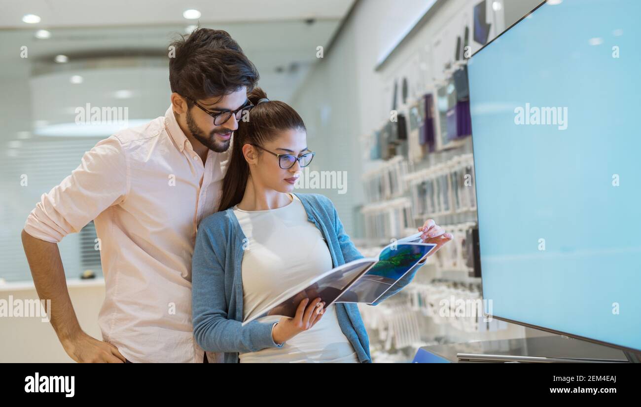 Giovane hipster elegante coppia curiosa lettura specifica di fronte al grande Flat TV nel negozio di tecnologia. Foto Stock