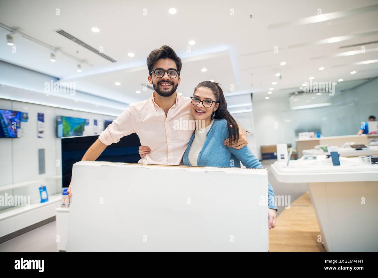 Giovane coppia di amore felice in piedi abbracciata con grande cartone animato dopo l'acquisto di un nuovo prodotto elettronico nel negozio di tecnologia. Foto Stock