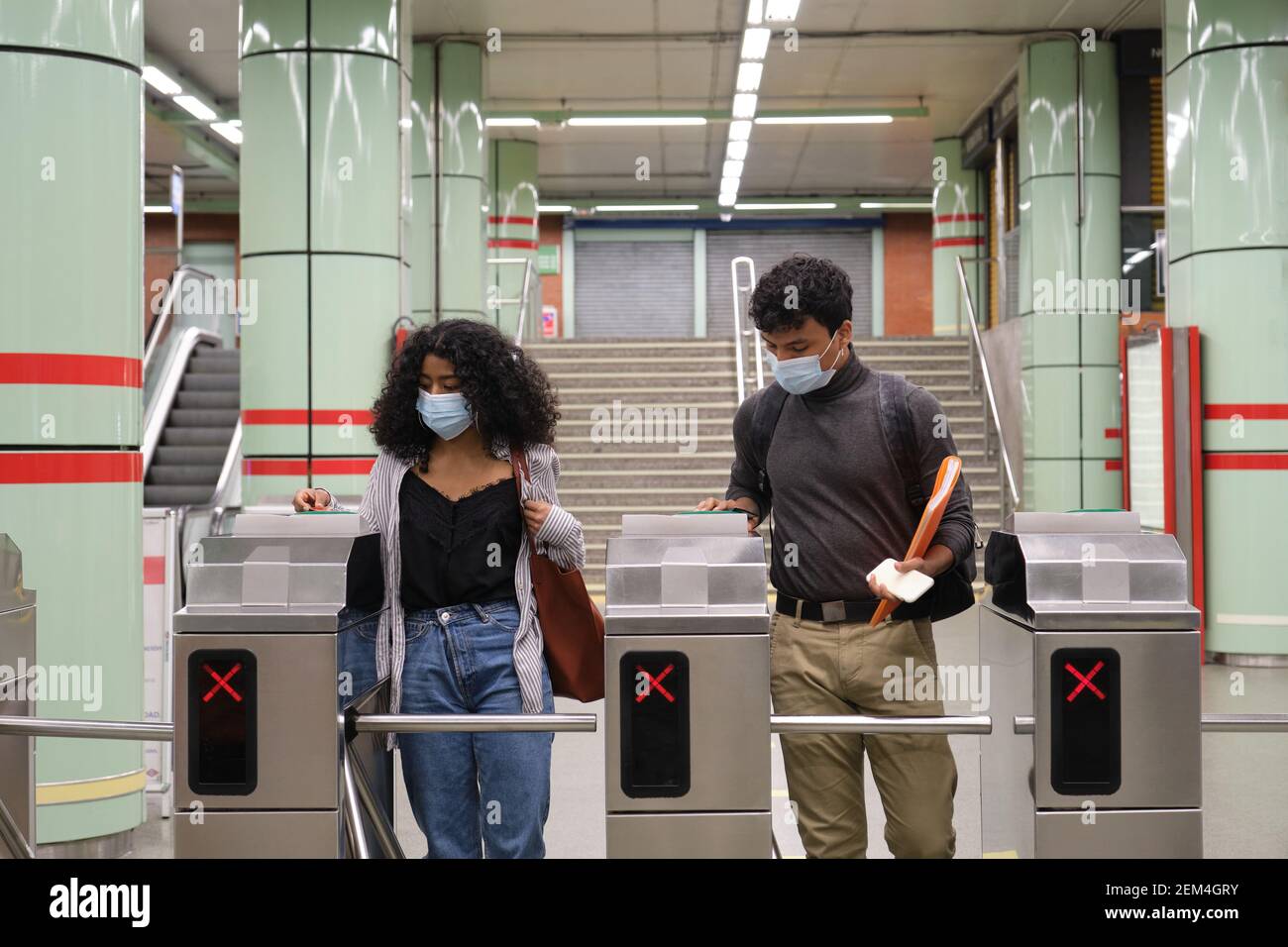 Giovane coppia latina con maschera protettiva che cammina attraverso il tornello della metropolitana alla stazione ferroviaria o della metropolitana. Nuovo normale ai trasporti pubblici. Foto Stock