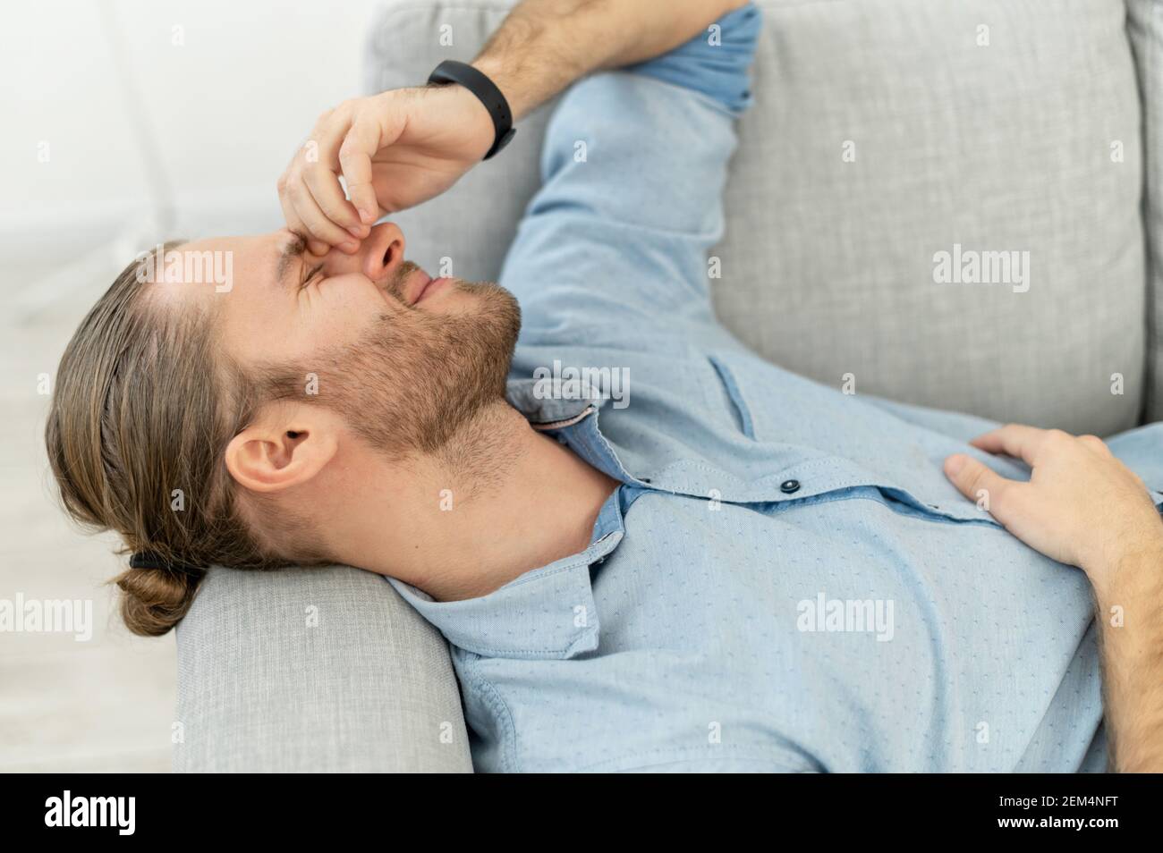 Uomo attraente con capelli marrone chiaro tirati indietro, barba, sdraiato sul divano grigio accogliente, con sorriso di piacere, tenendo il braccio sul viso, ricordando questo fantastico giorno, è stato un lavoro atteso da tempo, dopo la data Foto Stock