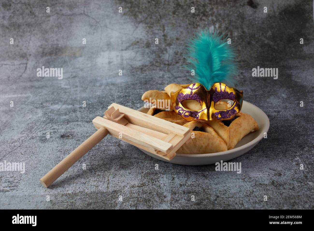 Concetto di celebrazione Purim con biscotti hamantashen, maschera Purim e noisemaker giocattolo su sfondo grigio Foto Stock