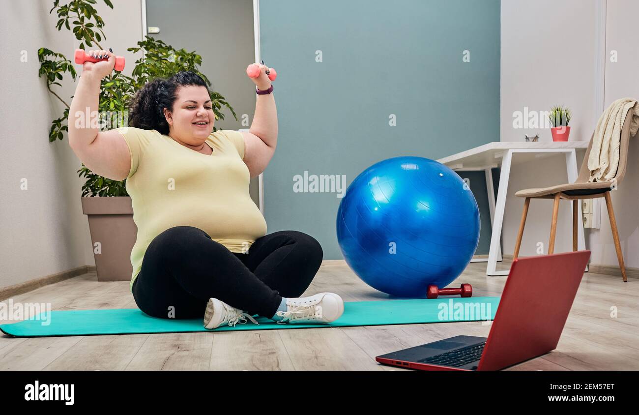 Donna in sovrappeso che fa esercizi a casa con personal trainer online per la perdita di peso usando i manubri Foto Stock