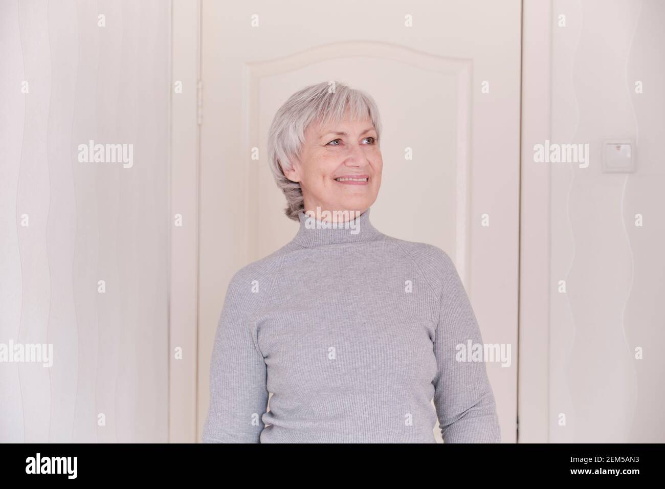 Un ritratto a vita di una donna caucasica più anziana sorridente con capelli corti grigi in un dolcevita grigio a casa. Foto Stock