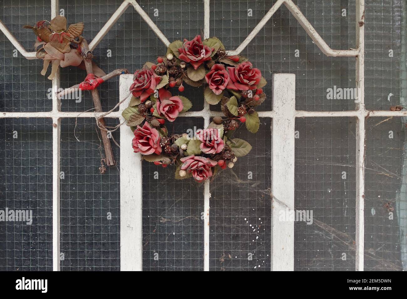 Decorazioni natalizie ornamenti vintage ricoperti di ciottoli sulla porta della casa abbandonata. Corona di plastica rosa fiori rossi cupido con lira e cros di legno Foto Stock