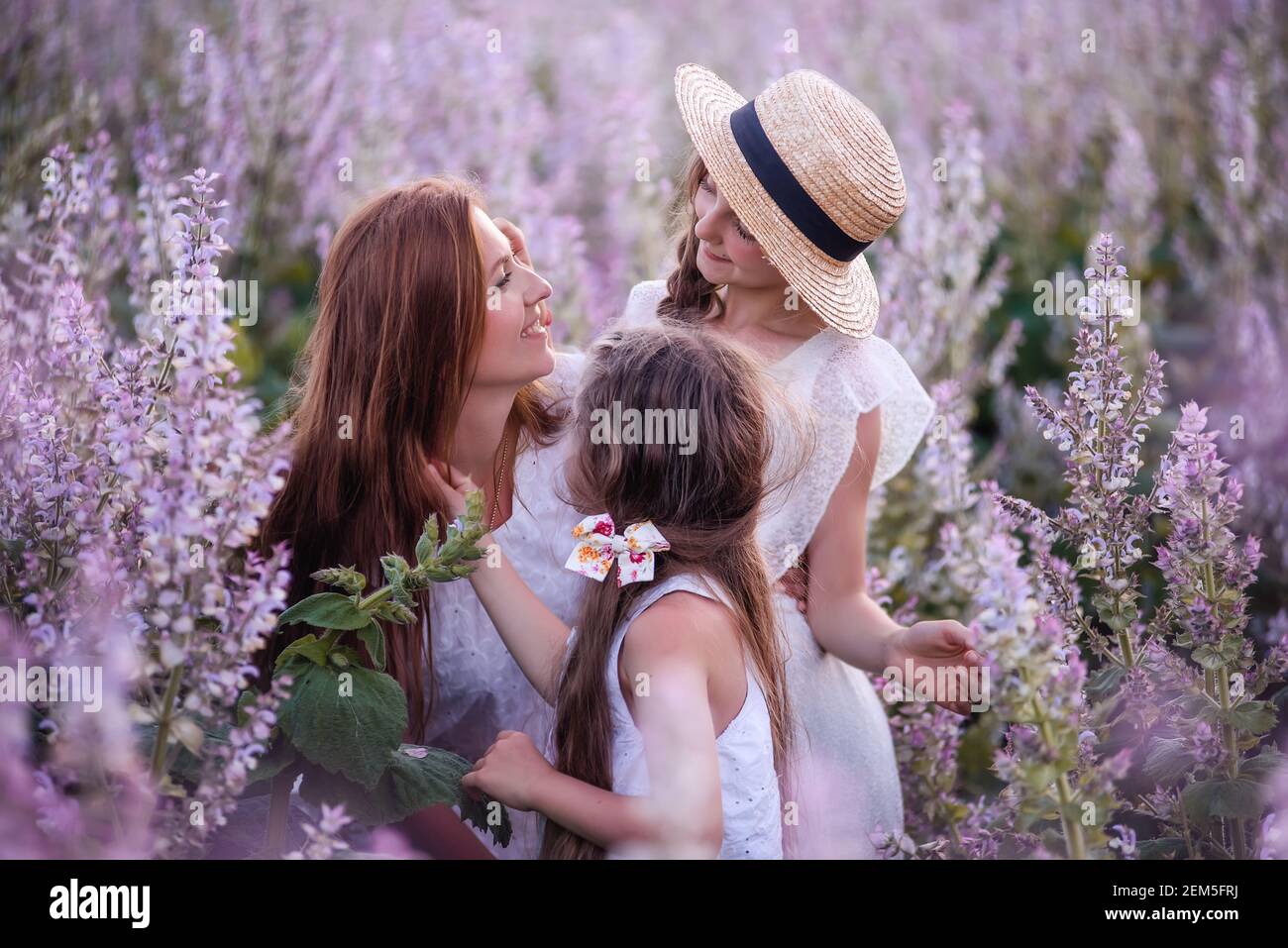 La giovane madre abbraccia due figlie nel campo di salvia in fiore viola. Le ragazze si abbracciano insieme, ridono, Buon divertimento. Vestito bianco con cappelli di paglia. In viaggio Foto Stock