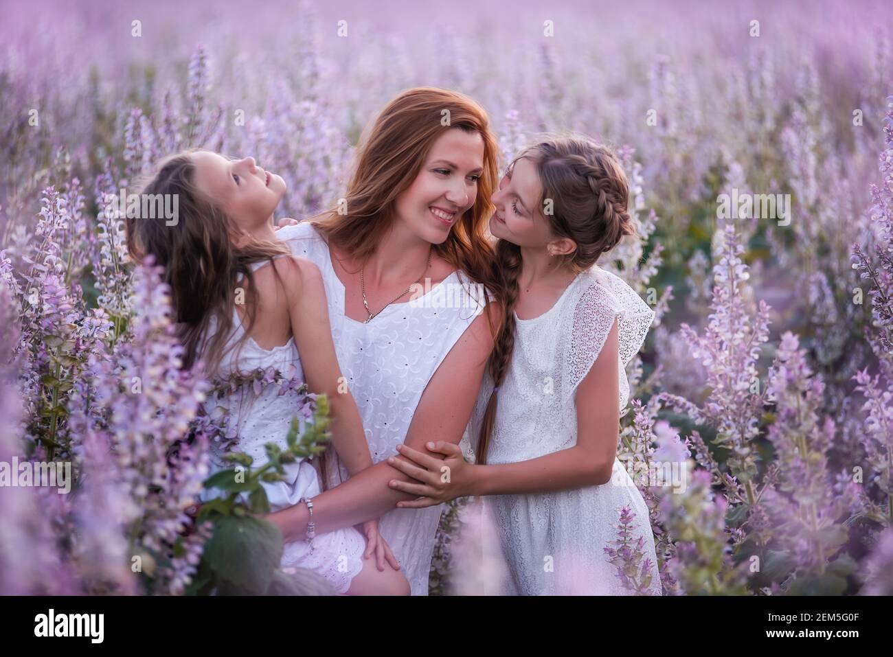 La giovane madre abbraccia due figlie nel campo di salvia in fiore viola. Le ragazze si abbracciano insieme, ridono, Buon divertimento. Vestito bianco con cappelli di paglia. In viaggio Foto Stock