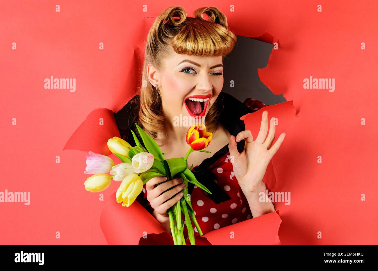 Winking donna con tulipani mostra segno ok. Sorridente Pinup ragazza che guarda attraverso la carta. Donna felice con bouquet di fiori. Foto Stock