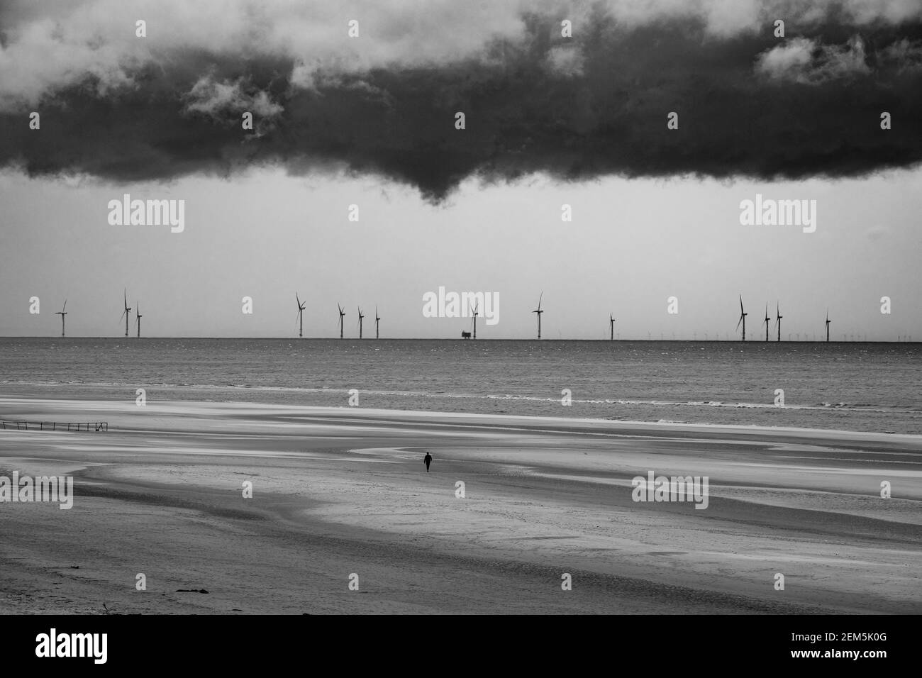 Un uomo solista cammina su una spiaggia vuota mentre le nubi tempestose si riuniscono sopra una fattoria eolica offshore. Foto Stock