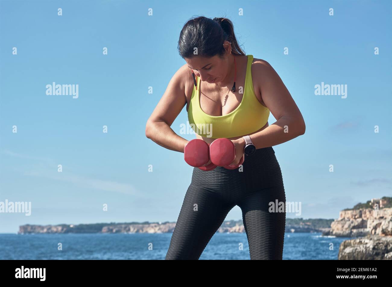 Donna latina di mezza età, con top giallo, guin nero, con manubri, ginnastica, riscaldato per addestramento, bruciare calorie, mantenersi in forma, all'aperto dal mare, Foto Stock