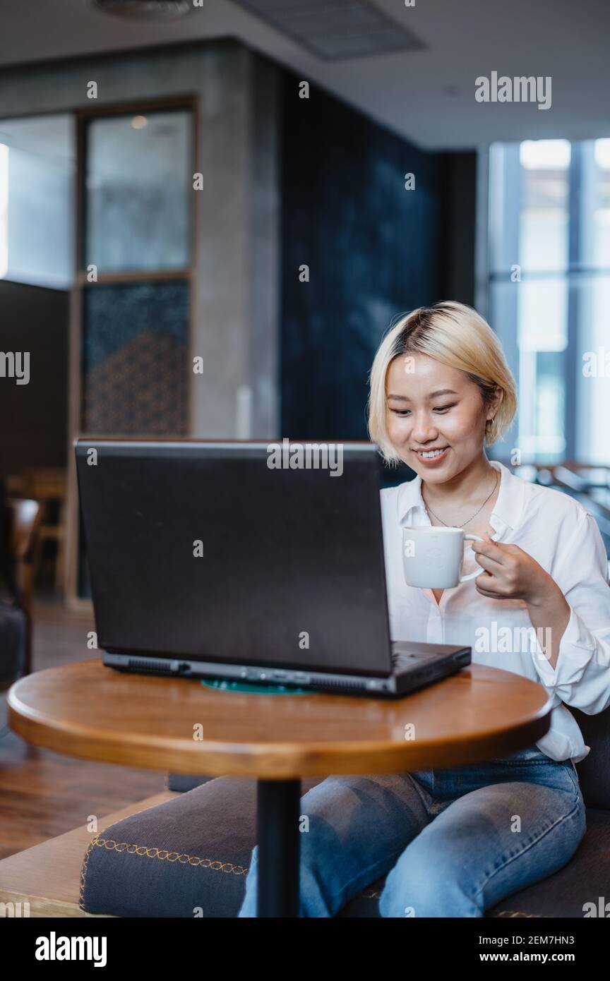 Giovane bella donna asiatica che lavora sul suo computer portatile in un bar, mentre beve caffè. Foto Stock