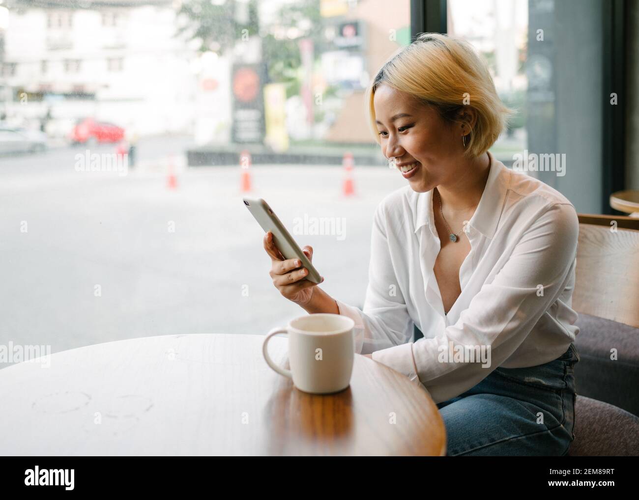 Giovane bella donna asiatica seduta in un caffè' vicino a una grande finestra, guardando il suo telefono sorridente. Foto Stock