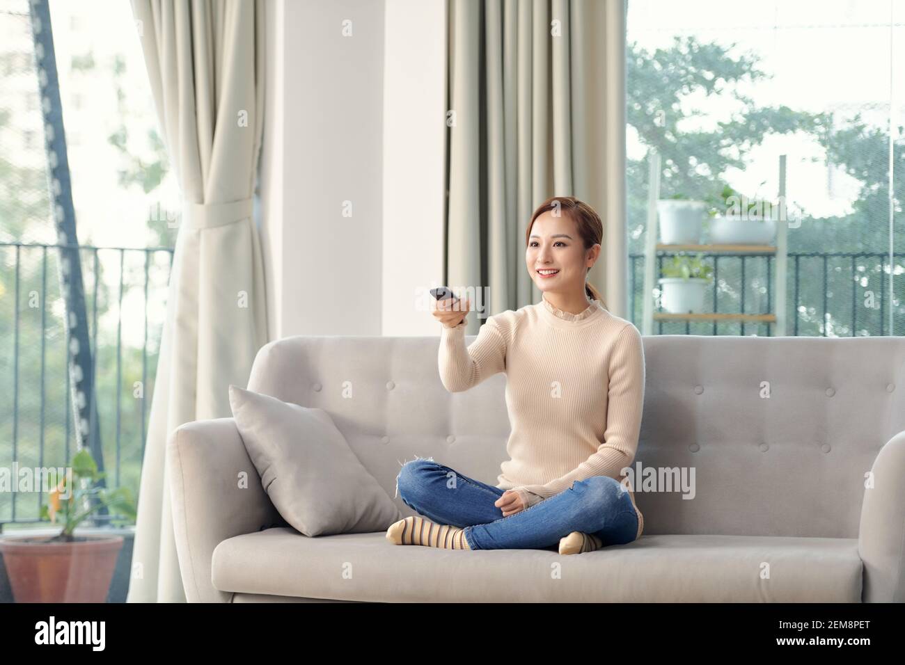 Foto di felice giovane donna seduta sul divano di casa. Guardando la telecamera tenendo premuto il telecomando per guardare la TV. Foto Stock