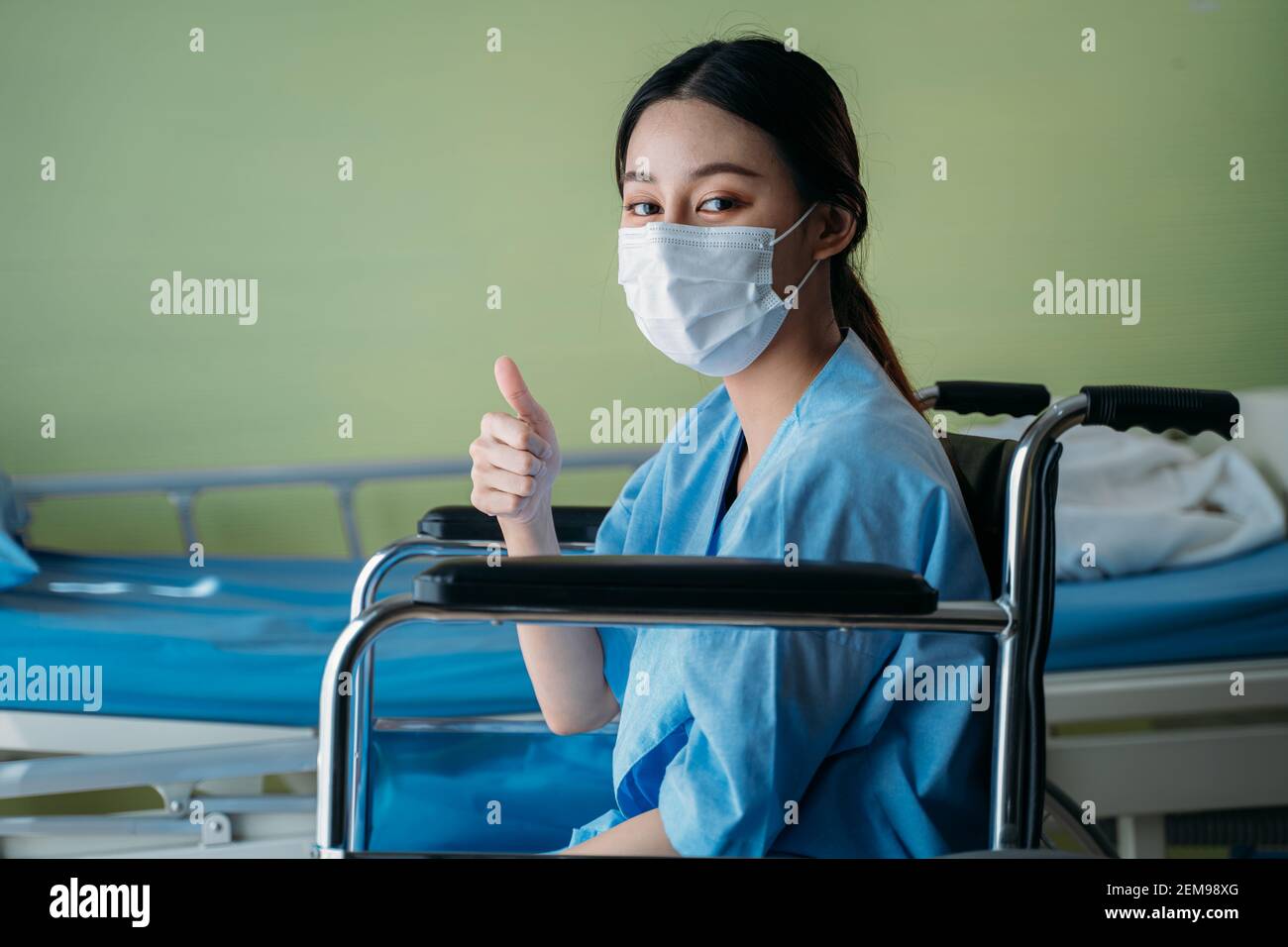 Ritratto di una giovane paziente asiatica sorridente con intervento chirurgico maschera seduta su sedia a rotelle nella stanza dell'ospedale che mostra i pollici verso l'alto gesti e segno sulla fotocamera Foto Stock