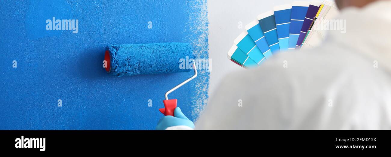 L'artigiano tiene il rullo e una tavolozza di colori e dipinge bianco blu parete Foto Stock