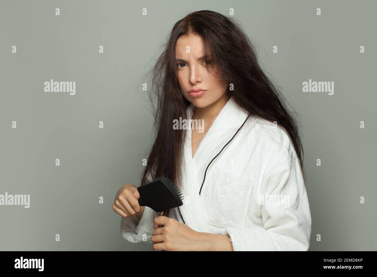 Donna bruna infelice che combatte i capelli aggrovigliati su sfondo bianco. Cura dei capelli e concetto di problema dei capelli Foto Stock