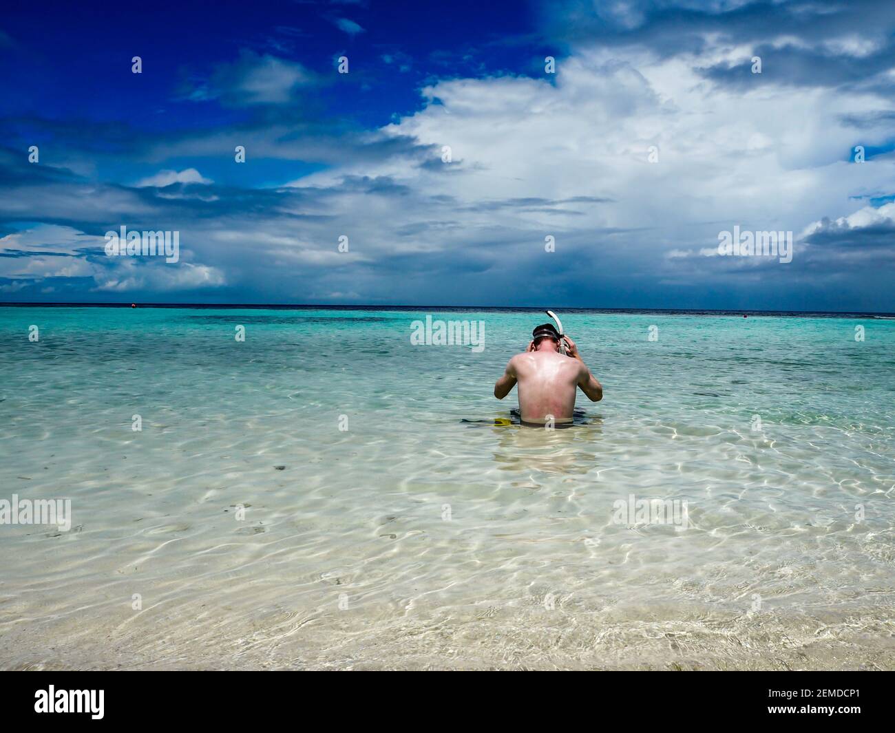 Giovane uomo seduto in acque poco profonde e limpide e che si mette su attrezzatura da snorkeling Foto Stock