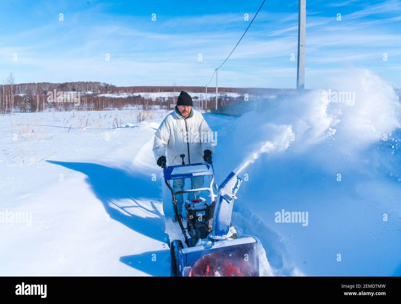 Un uomo in una giacca bianca rimuove la neve da a. strada rurale con un snowblower in inverno dopo una nevicata Foto Stock