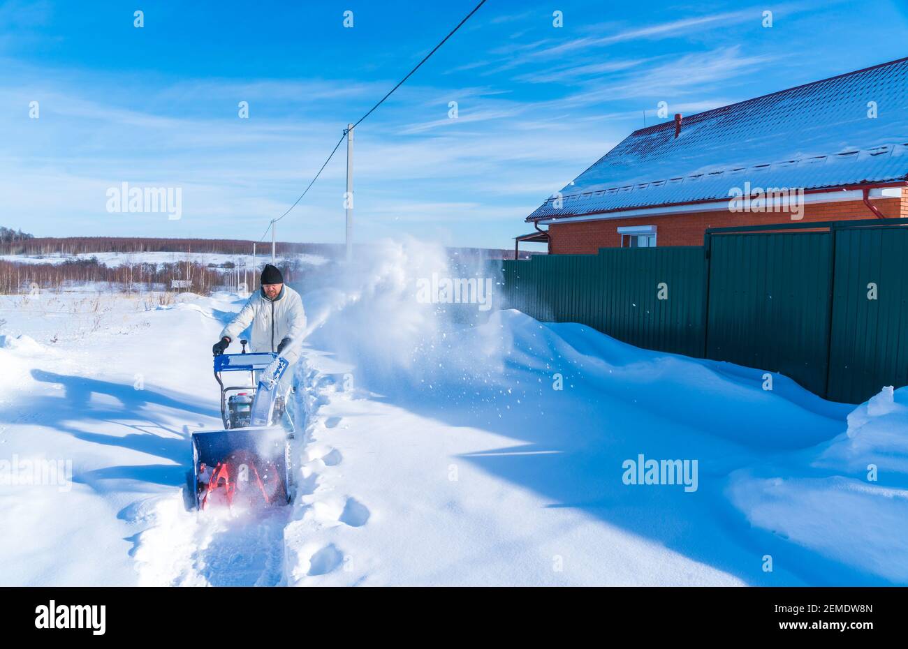 Un uomo in una giacca bianca rimuove la neve da a. strada rurale con un snowblower in inverno dopo una nevicata Foto Stock
