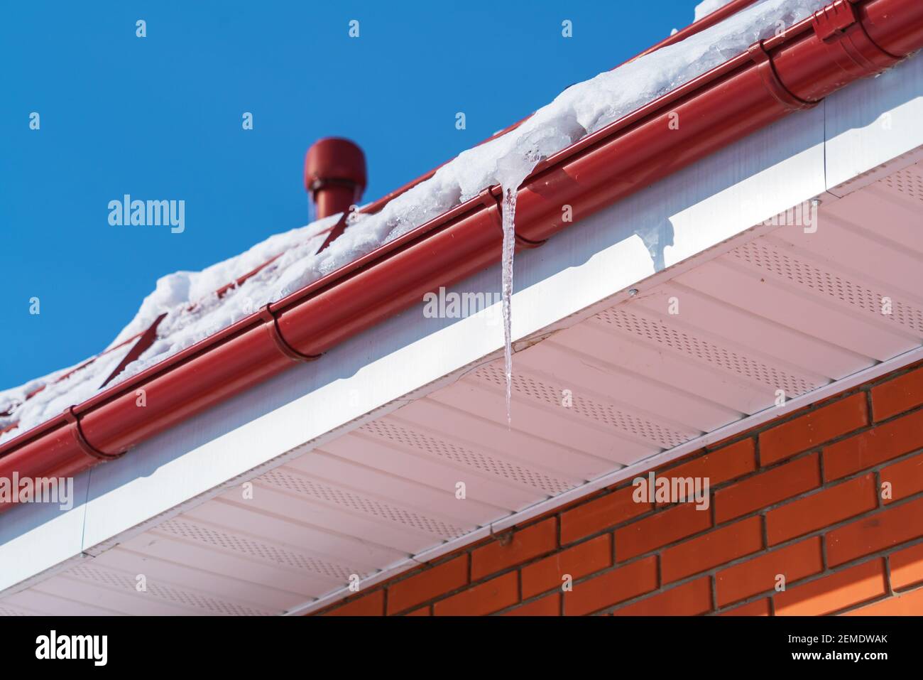 Un grande icicolo trasparente contro un cielo blu chiaro è sospeso da un tubo di scarico rosso in inverno alla vigilia di molla Foto Stock