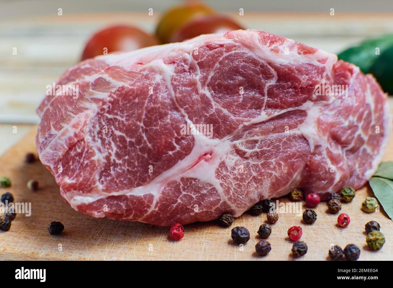 Un pezzo di carne cruda con grani di pepe e foglie di alloro su un tagliere. Sullo sfondo verdure fresche - pomodori e cetrioli Foto Stock