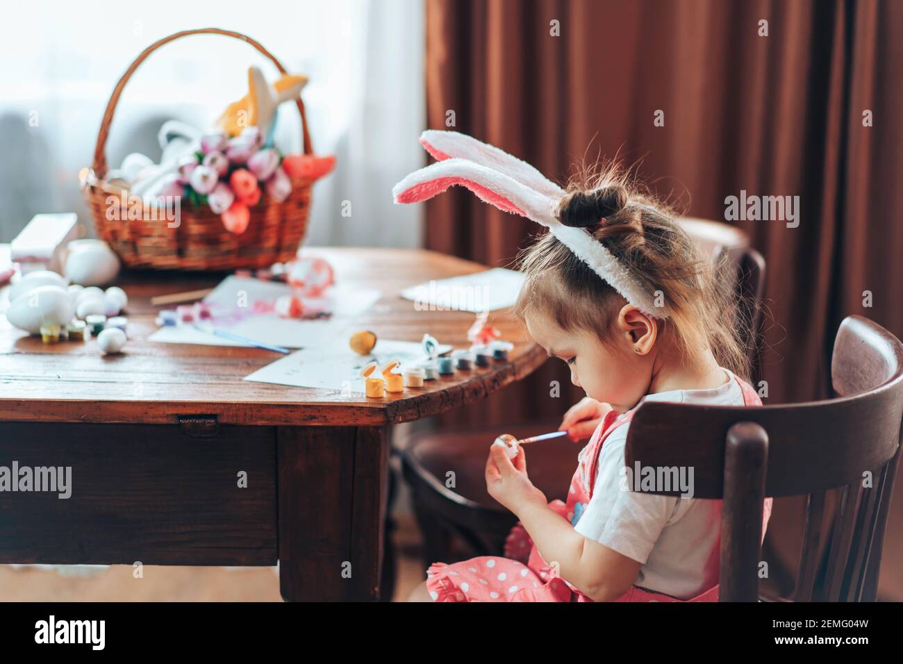 La ragazza sta dipingendo le uova di Pasqua per la vacanza ad un tavolo di legno. I bambini hanno orecchie conigliate sulle loro teste. Preparazione delle decorazioni pasquali per la vacanza. Foto Stock