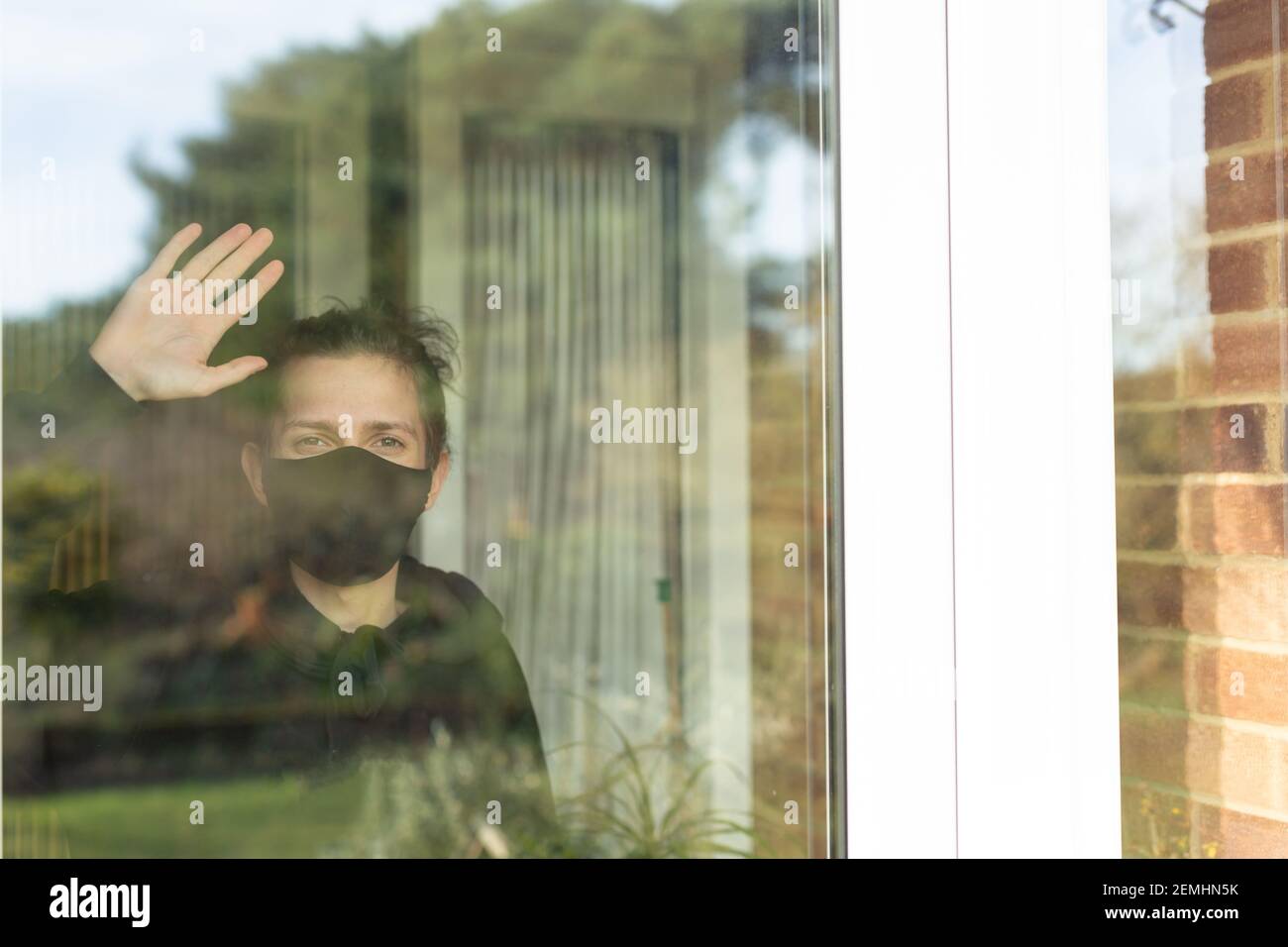 Un giovane uomo con una maschera facciale che guarda e sventola fuori la finestra durante il suo auto-isolamento a casa. Covid fine del concetto di libertà di blocco Foto Stock