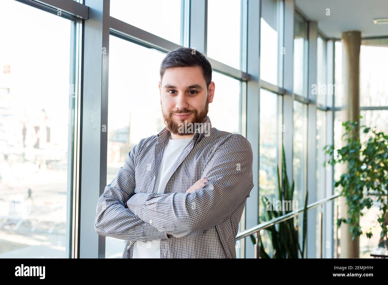 uomo caucasico sicuro in piedi vicino a tutta la finestra in ufficio con le piante. Il giovane bearded riuscito ha attraversato le braccia. Bell'uomo d'affari intelligente Foto Stock