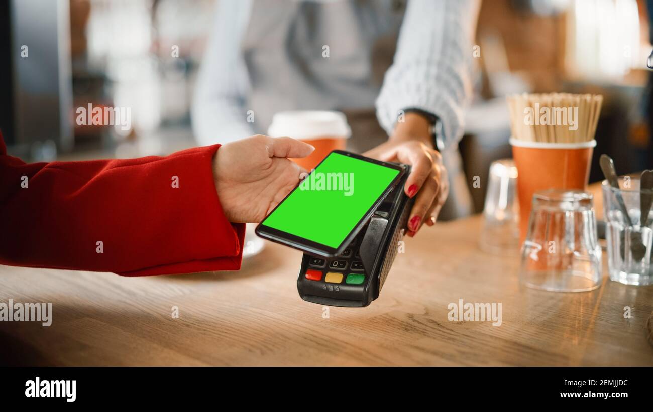 Primo piano di una mano femminile con cui tenere uno smartphone Modello schermo verde con tecnologia di pagamento NFC utilizzata per Pagando per il caffè da portare via in un Foto Stock