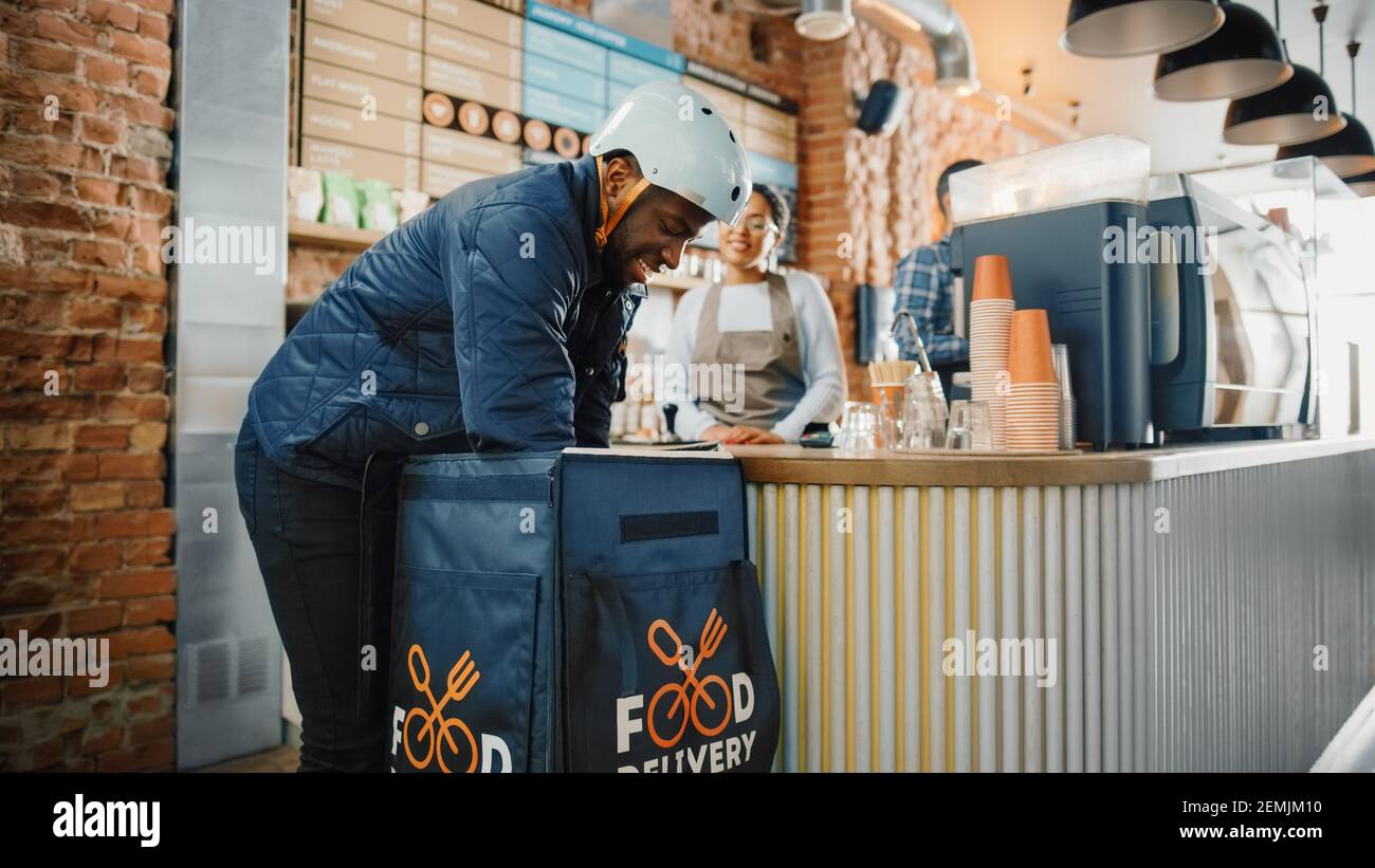 Beautiful Happy Latin Barista serve ordine a un corriere di consegna cibo raccolta sacchetto di carta con pasticcini da un ristorante Cafe. Delivery Guy mette Foto Stock