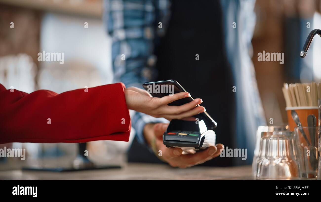 Primo piano di una mano femminile tenere uno smartphone con una tecnologia di pagamento NFC utilizzata per pagare il caffè da portare via in un caffè. Il cliente utilizza Mobile Foto Stock