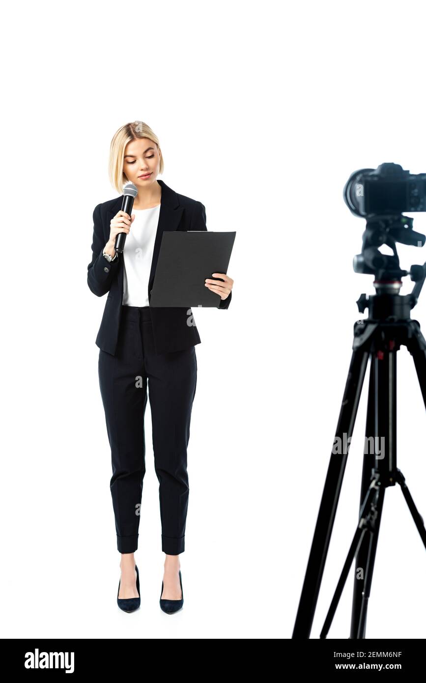 vista a tutta lunghezza dell'ancoraggio di notizie bionde in tuta nera vicino alla fotocamera digitale su un primo piano bianco sfocato Foto Stock