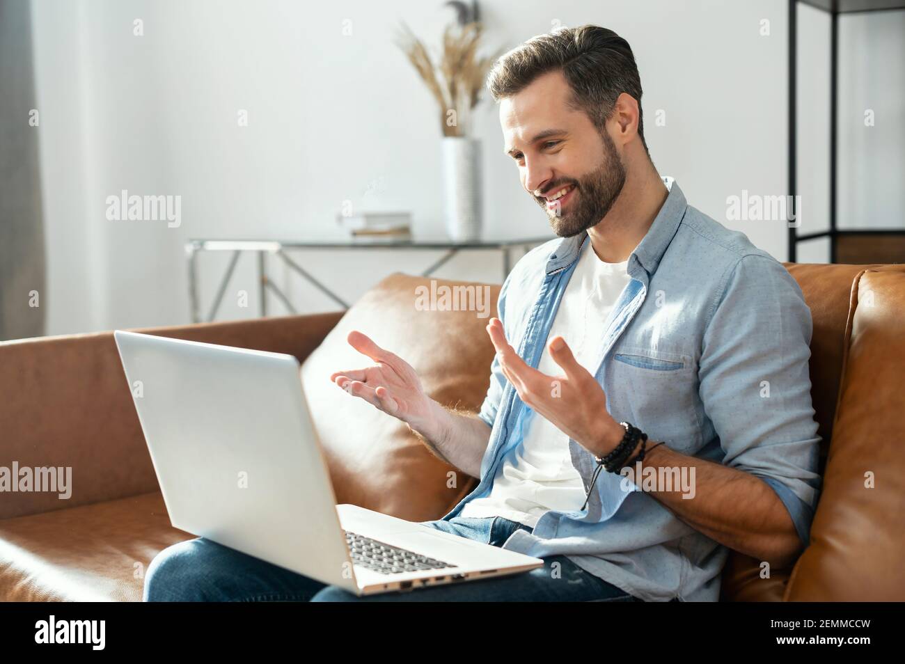 Sorridente giovane uomo hipster sopportato seduto sul divano con il laptop in grembo, flirtare, utilizzando l'app di datazione, videochiamata ragazza online, gesturing e parlare, blogger streaming la sua vita quotidiana Foto Stock