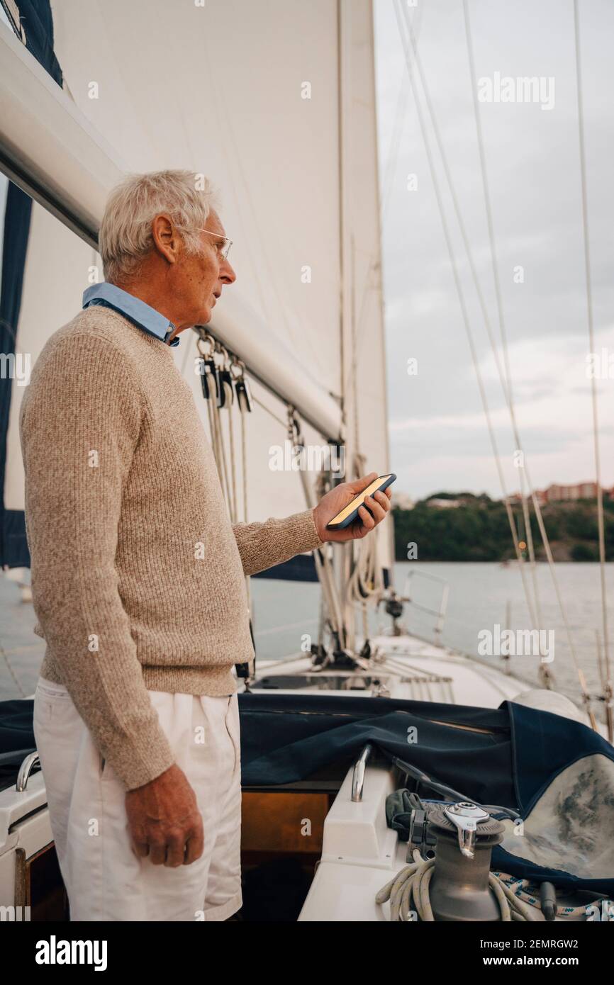 Uomo anziano che segue la direzione attraverso lo smartphone mentre si è in piedi barca durante il tramonto Foto Stock