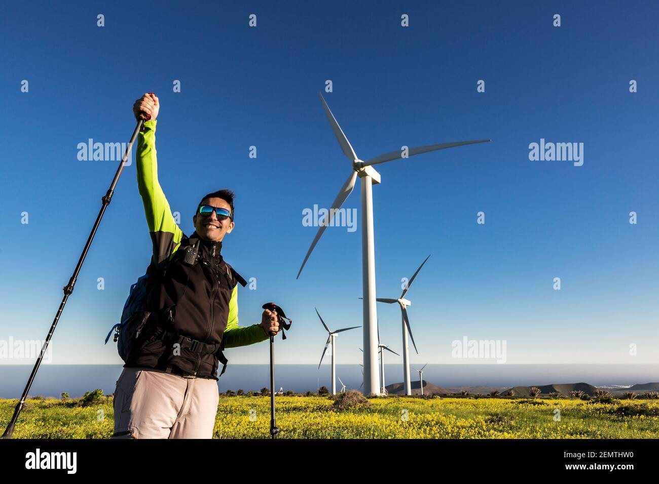 Sorridente escursionista maschile con bastoni da trekking in piedi con la mano sollevata e celebrando la vittoria sullo sfondo di mulini a vento in campo su Lanzarote Foto Stock