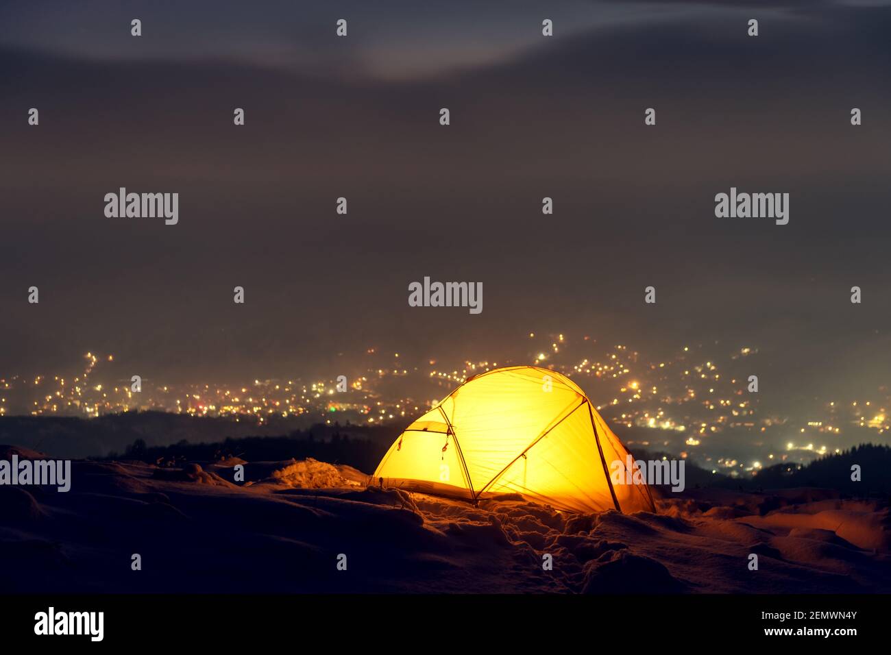 Tenda gialla illuminata dall'interno sullo sfondo di luci della città incandescenti nella nebbia. Incredibile paesaggio innevato. Campo turistico in montagna d'inverno. Concetto di viaggio Foto Stock