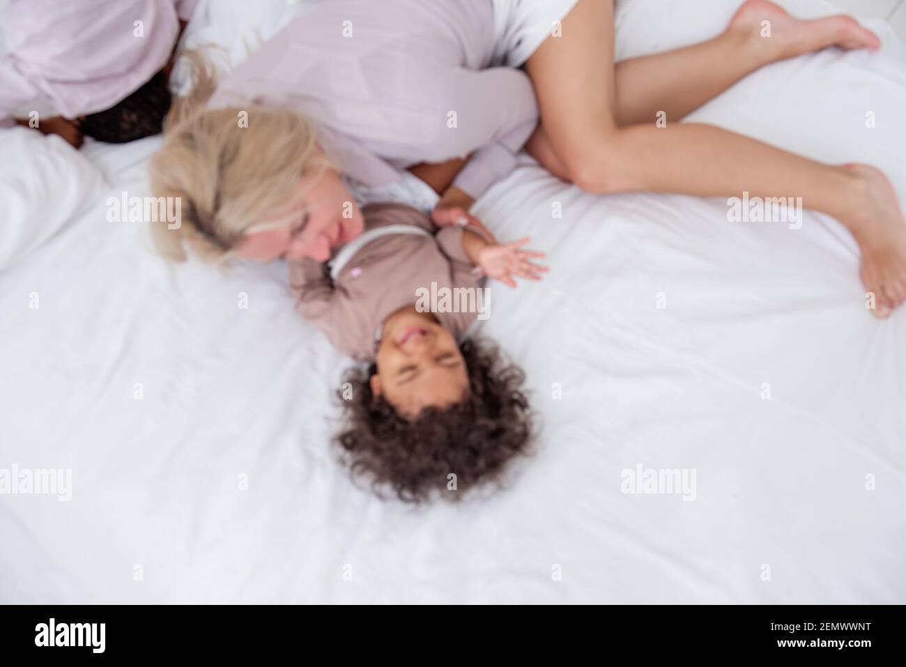 Defocused giovane madre bionda caucasica solletico la sua piccola figlia afroamericana. Sdraiati sul letto bianco, divertendoti, con vista dall'alto. Le ragazze ridono, abbraccio, il Foto Stock