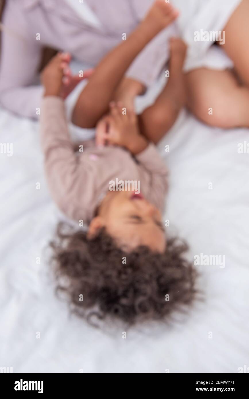 Defocused giovane madre bionda caucasica solletico la sua piccola figlia afroamericana. Sdraiati sul letto bianco, divertendoti, con vista dall'alto. Le ragazze ridono, abbraccio, il Foto Stock