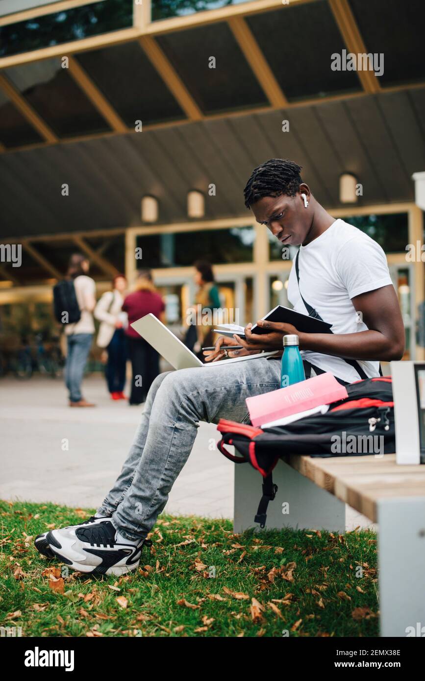 Alunno africano maschio libro di lettura mentre si siede su panca dentro campus universitario Foto Stock