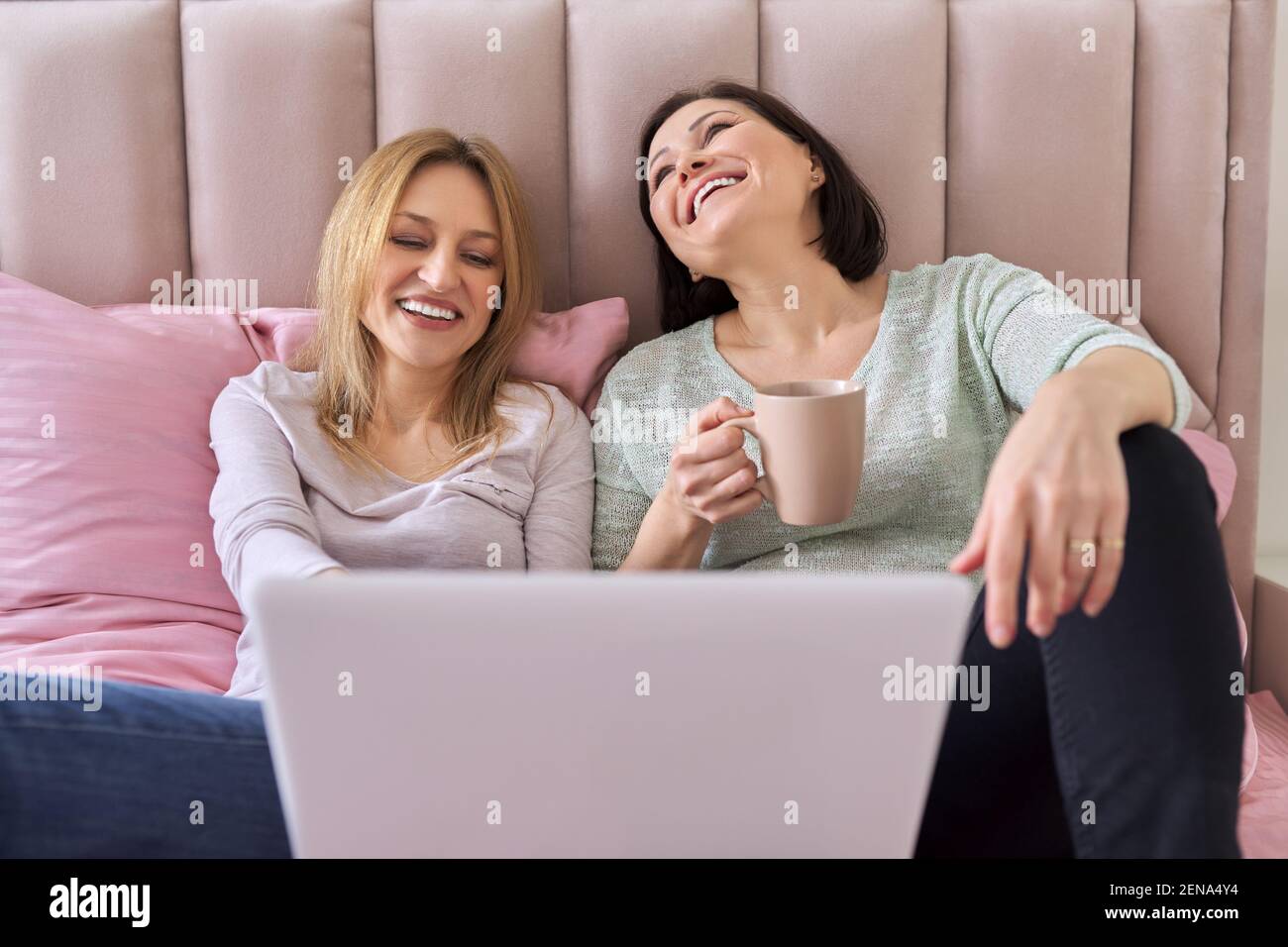 Due donne mature che guardano un notebook seduto a casa letto Foto Stock