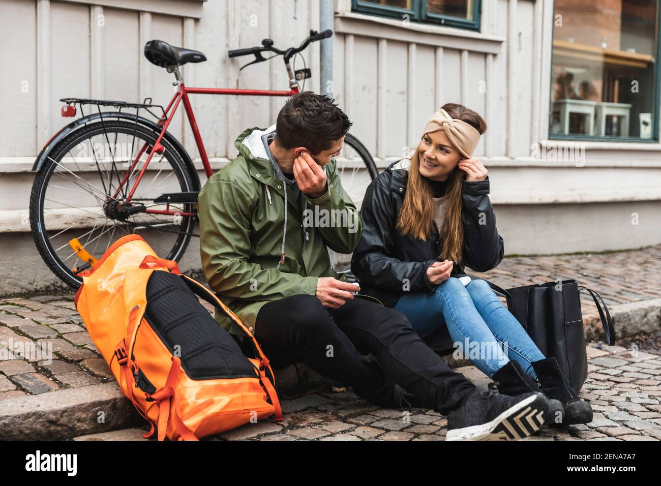 Turisti di sesso maschile e femminile che indossano cuffie intrauricolari mentre sono seduti sentiero in città Foto Stock