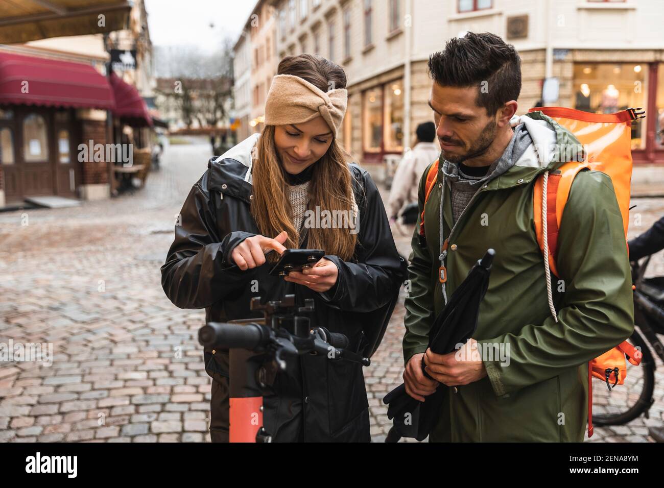 Coppia eterosessuale che usa lo smartphone mentre in piedi scooter elettrico push sulla strada in città Foto Stock